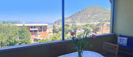 A balcony with a table and chairs, a vase with flowers, and a view of a cityscape and mountains.