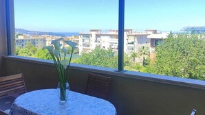 Un balcone con tavolo e sedie, un vaso con fiori e vista su edifici e vegetazione.