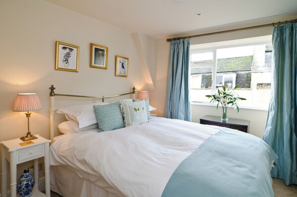 A neatly made bed with white and blue bedding, a bedside table with a lamp, and a window with curtains.