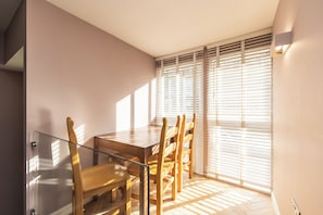 Un comedor moderno con una mesa de vidrio, sillas de madera y vistas a través de ventanas grandes con persianas.