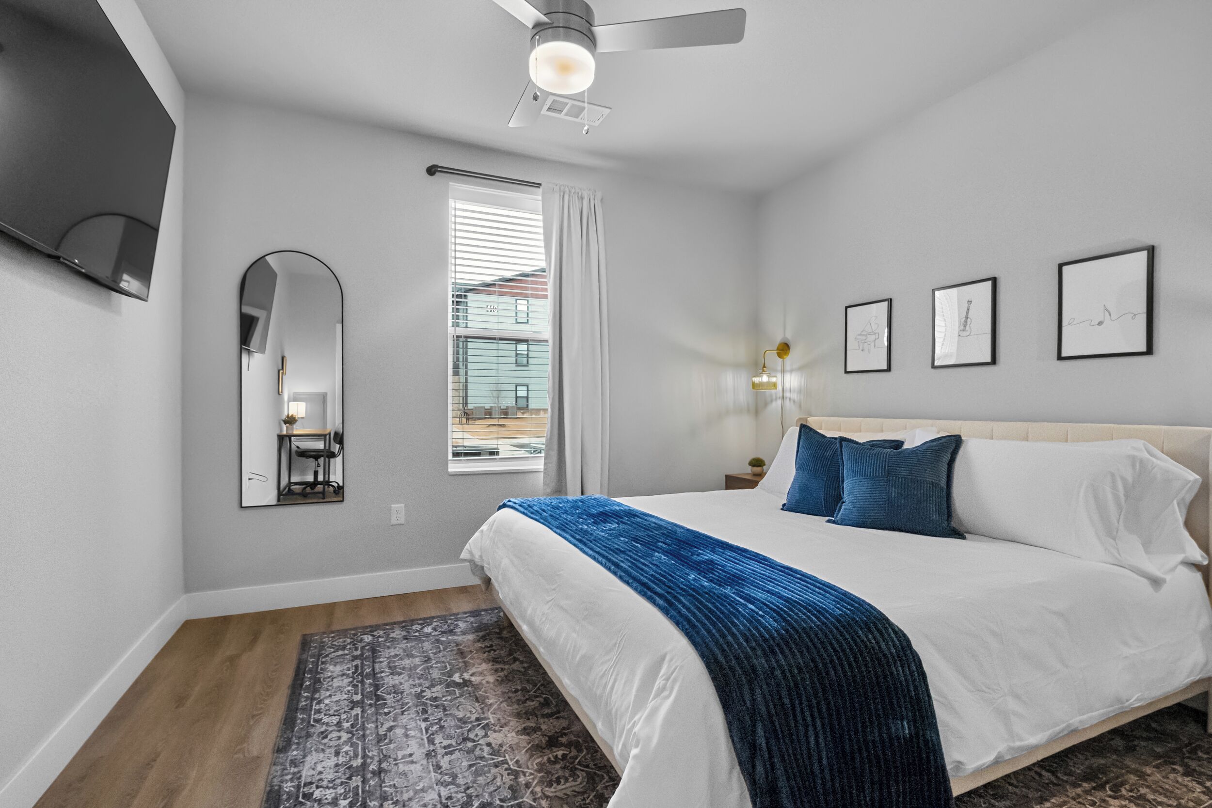 A bedroom with a bed, a ceiling fan, a window with curtains, a wall-mounted TV, and framed artwork.