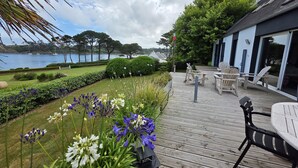 A wooden deck with outdoor furniture, a view of a lake, and a building with a flag. - Beautiful house with incredible sea view over Aber Wrach (Landéda)