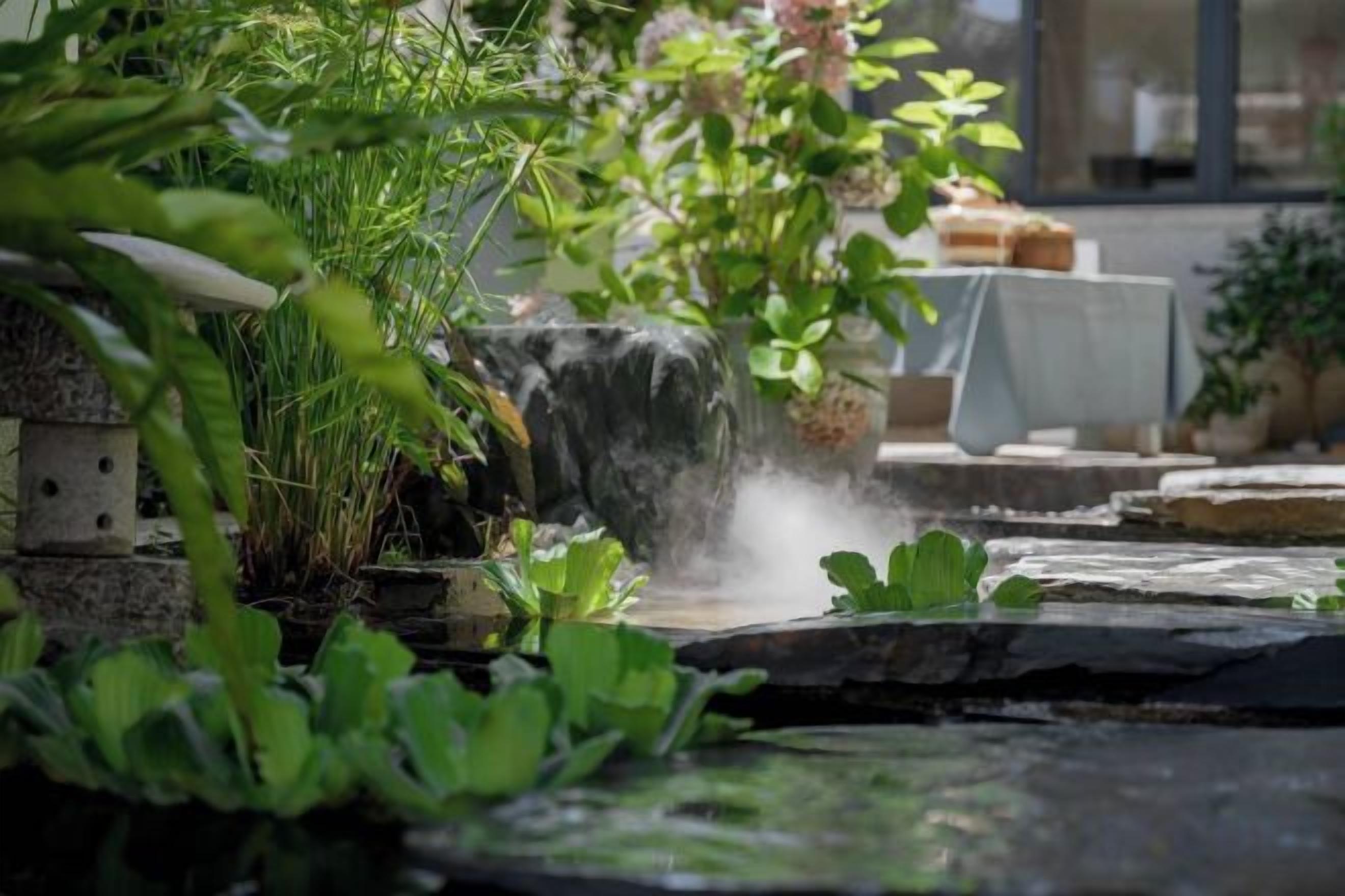A garden with a small waterfall, a table with chairs, and various plants.