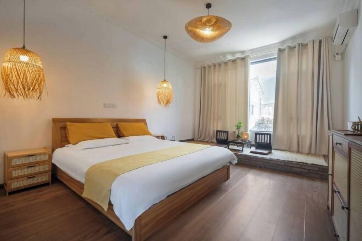Une chambre à coucher avec un lit en bois, deux lampes suspendues, une fenêtre avec des rideaux et une vue sur l’extérieur.