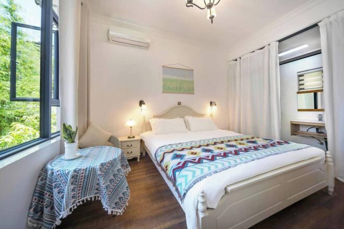 Une chambre à coucher avec un lit, des tables de chevet, une fenêtre avec des rideaux, une plante et un tableau au mur.