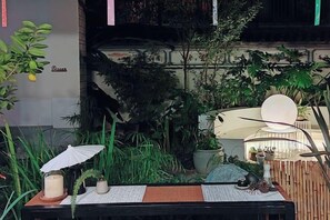 Une table dressée avec un menu, des bougies et un parasol décoratif, dans un jardin intérieur agrémenté de plantes en pot et d’une chaise suspendue.