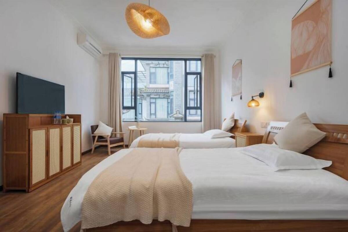 Une chambre d’hôtel avec deux lits, une armoire en bois, une télévision, une fenêtre donnant sur des immeubles et un luminaire suspendu.
