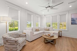 Uma sala de estar moderna com um sofá seccional branco, uma poltrona listrada, uma mesa de centro de madeira e janelas amplas com vista para vegetação.