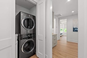 Un cuarto de lavado con dos lavadoras y secadoras apiladas, una puerta blanca y un pasillo que conduce a una habitación con ventana y un cuadro.