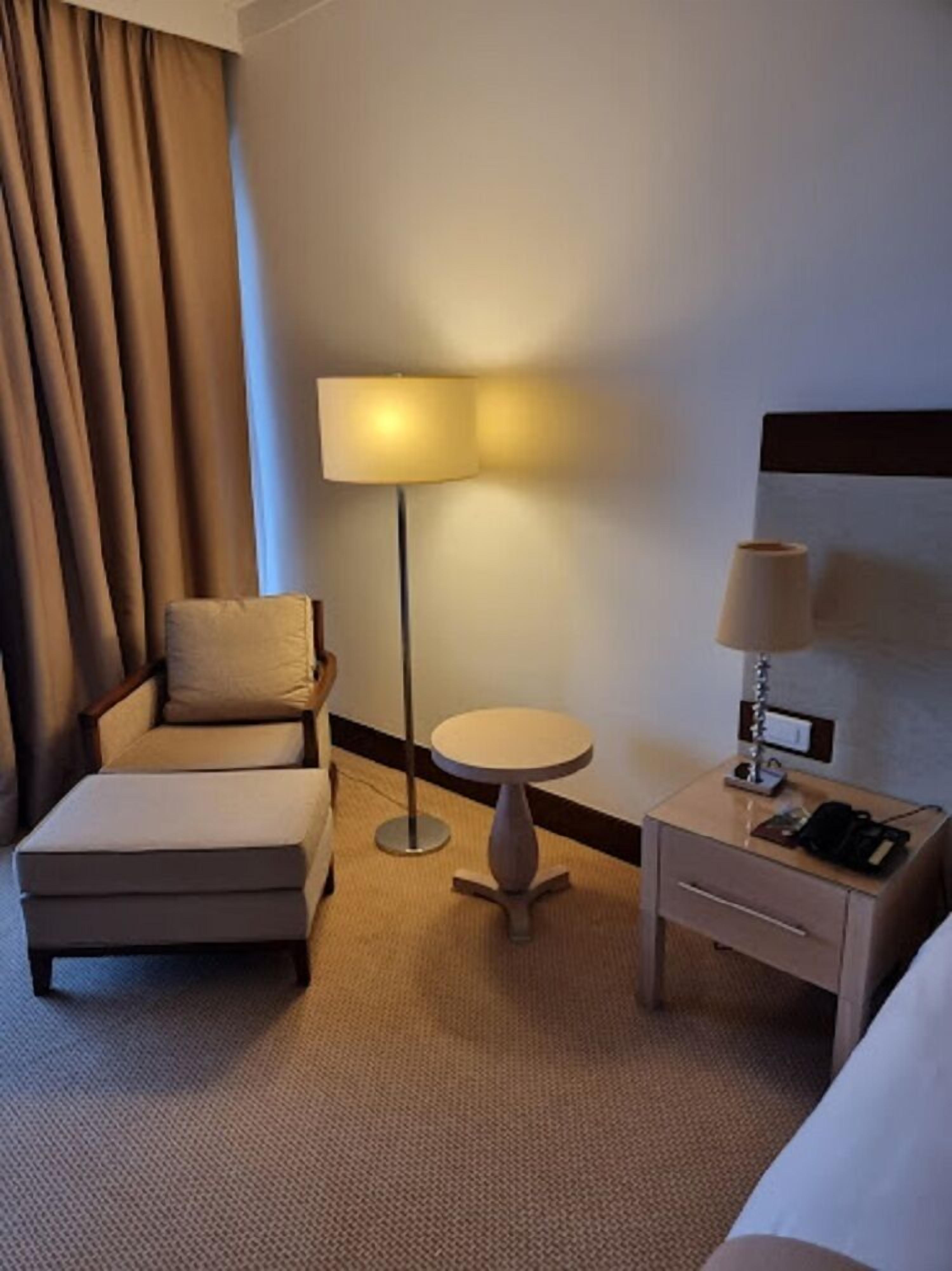 A hotel room with a bed, a chair, a small round table, a lamp, and a nightstand with a phone.