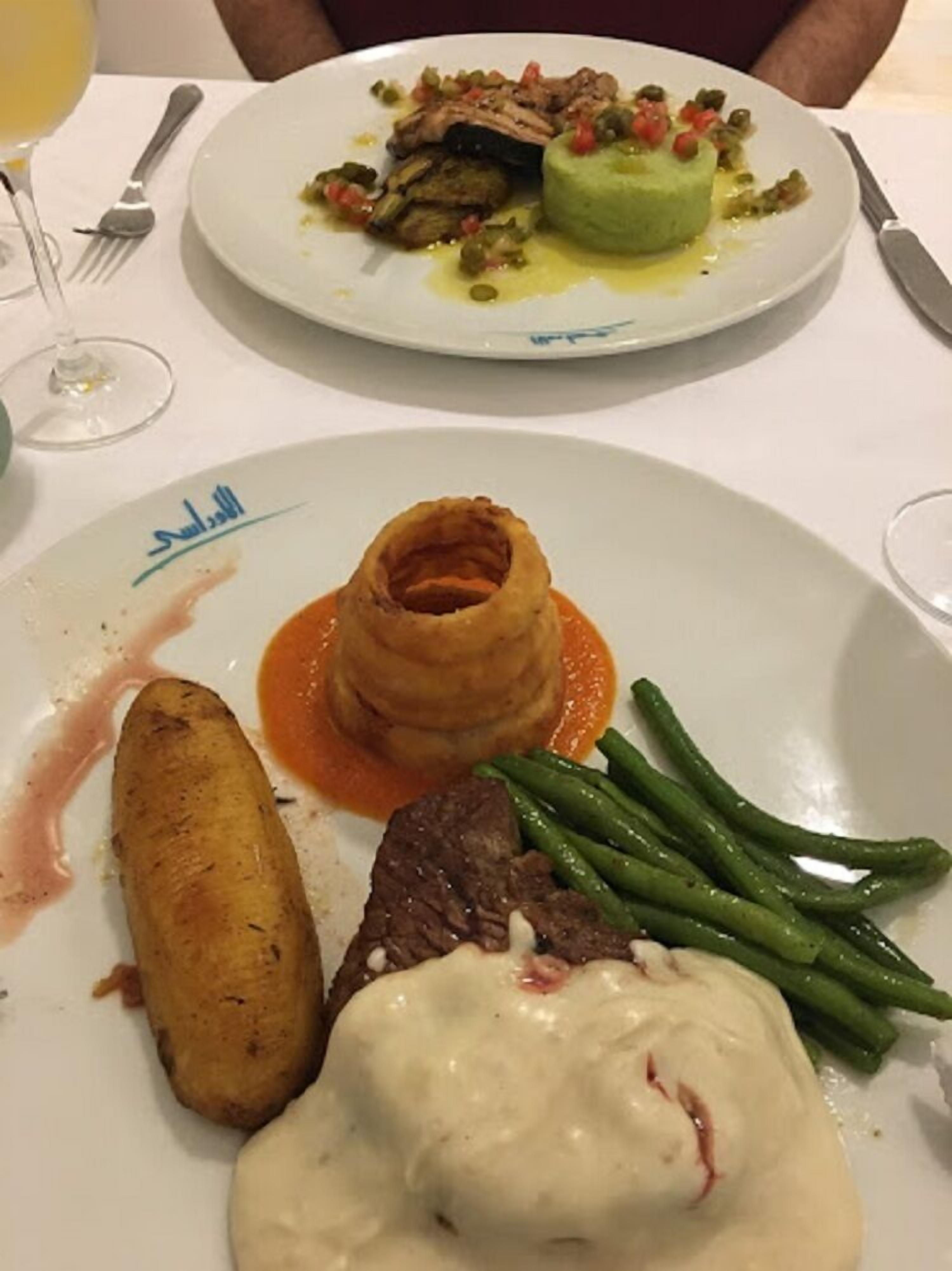 Een eettafel met twee borden eten, waarvan één een steak en groene bonen bevat en de ander verschillende groenten en een stuk vlees.