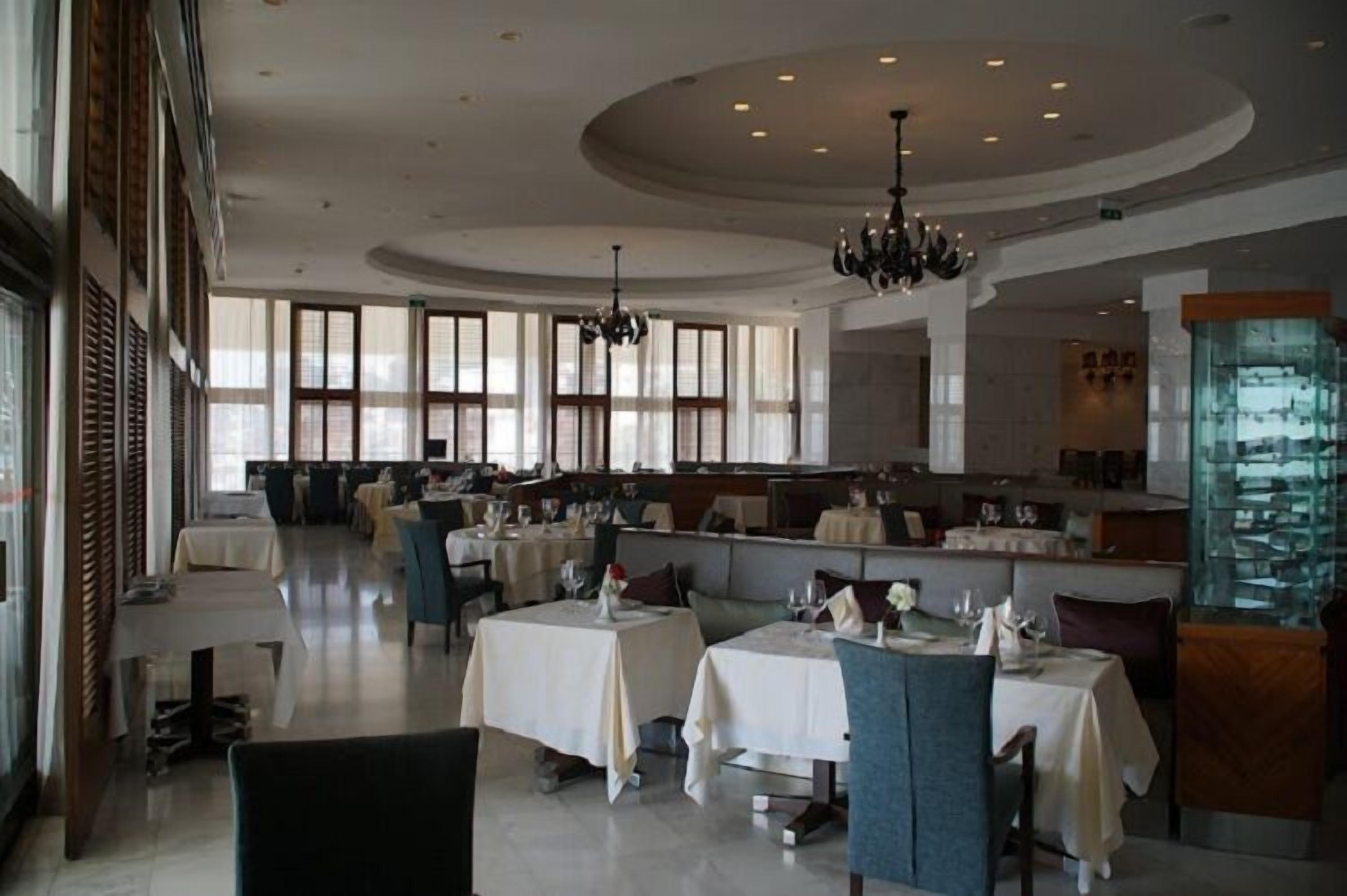Un ristorante spazioso con soffitti alti, ampie finestre e un'area bar centrale. I tavoli sono apparecchiati con tovaglie bianche e sedie.
