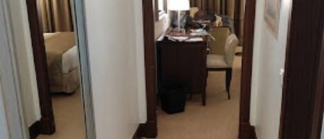 A hotel hallway with a carpeted floor, a large mirror on the left, a desk with a lamp, and a chair in the background.