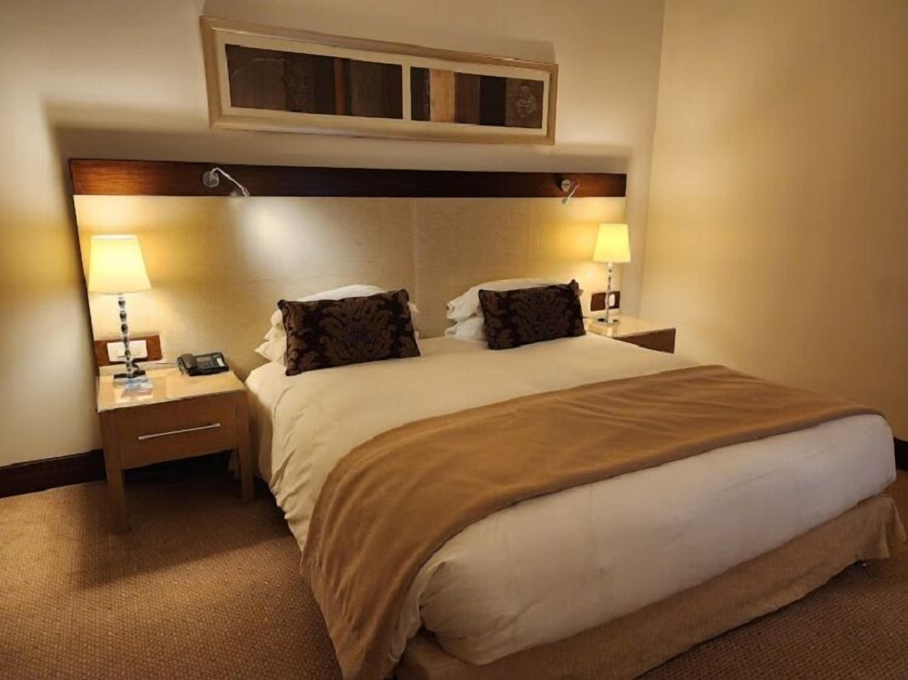 Habitación de hotel con una cama grande, dos mesitas de noche, lámparas y un cabecero con un panel decorativo.