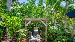 A wooden pergola structure in a lush garden with a paved pathway leading through it. - Paradise Pointe by Beach Boutique Rentals (Anna Maria)