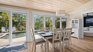 A modern kitchen with white cabinets, a dining area with a table and chairs, and a view of a swimming pool and greenery outside. - Paradise Pointe by Beach Boutique Rentals (Anna Maria)