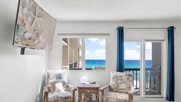Ein Zimmer mit zwei Sesseln, einem kleinen Tisch und einem Fernseher an der Wand. Es gibt ein großes Fenster mit Blick aufs Meer.