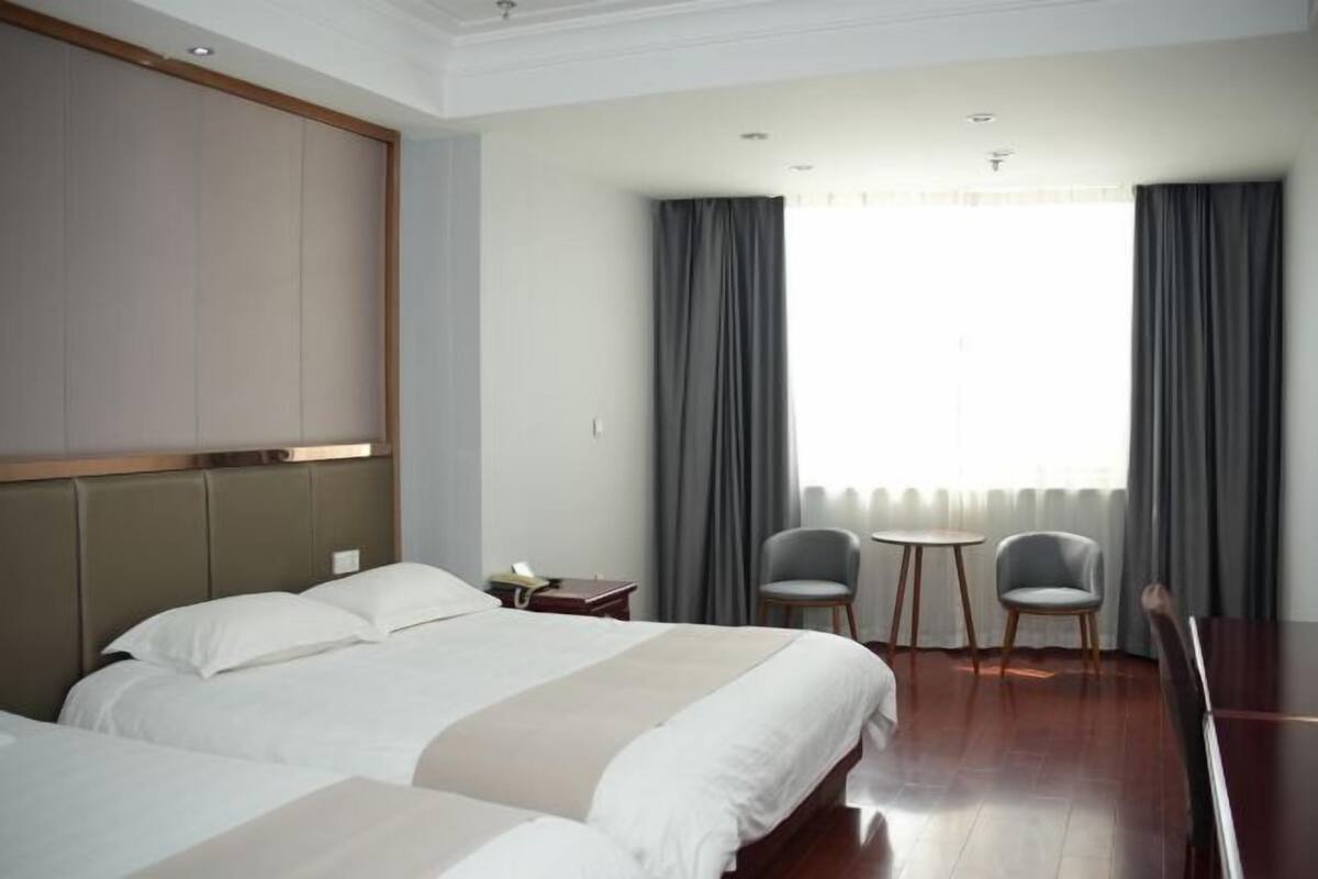 A hotel room with a large bed, two bedside tables, a small desk, two chairs, a round table, and a large window with curtains.