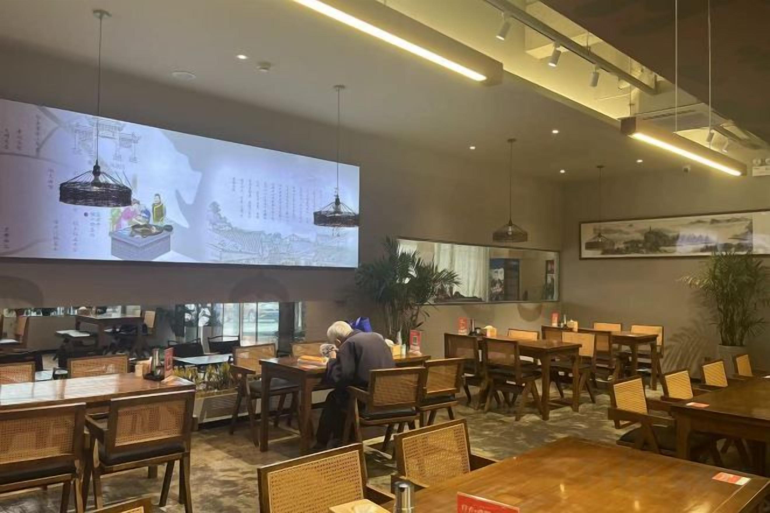 A restaurant with multiple tables and chairs, a large projection screen displaying menu items, and a waiter attending to a table.