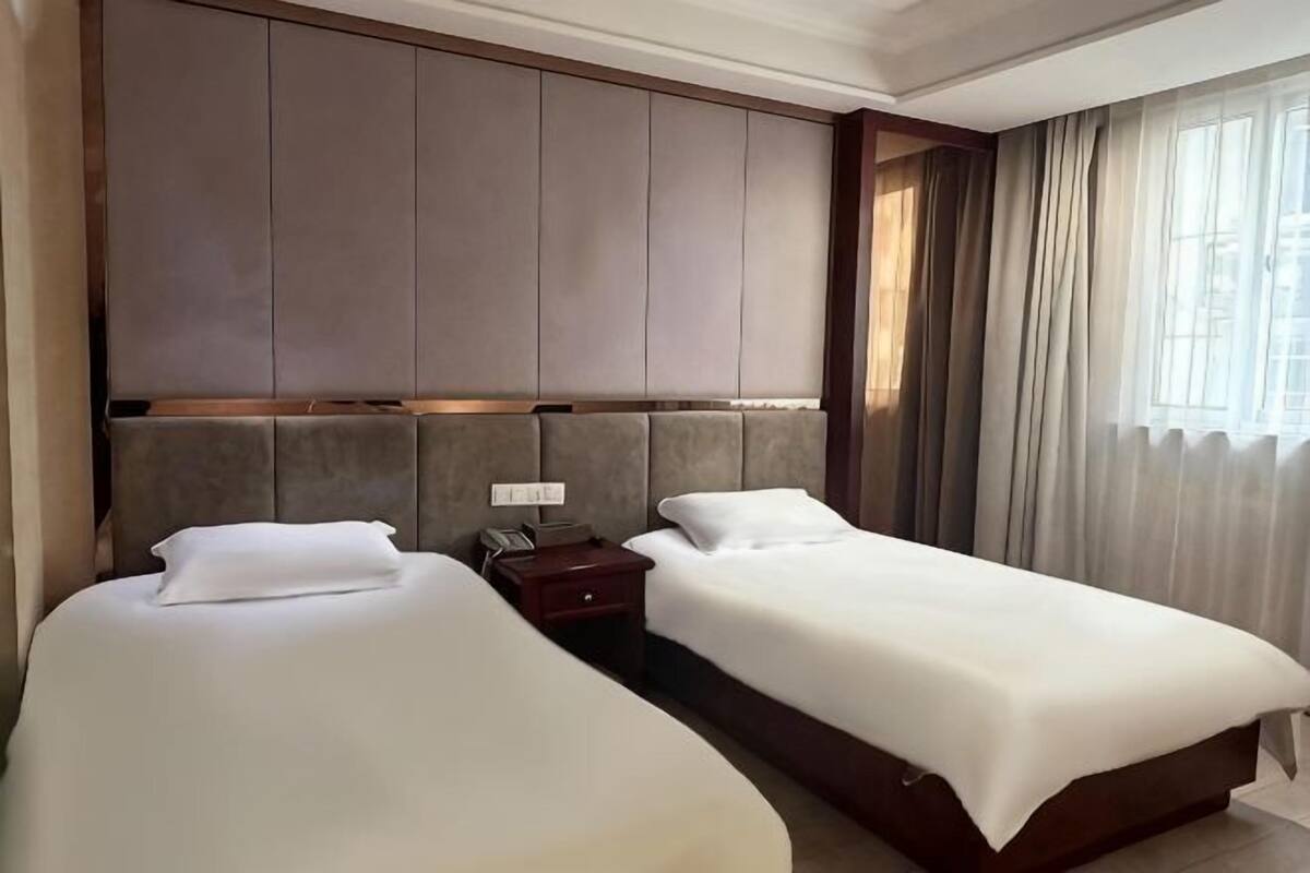 A hotel room with two beds, a large padded headboard, a bedside table with a phone, and a window with curtains.