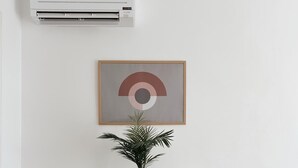 Et hvidt skab med et indrammet abstrakt kunstværk over, en potteplante på en flettet skammel, og en aircondition-enhed monteret på væggen.