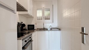 Et lille køkken med hvide skabe, en mikrobølgeovn, komfur og et vindue med rullegardiner.