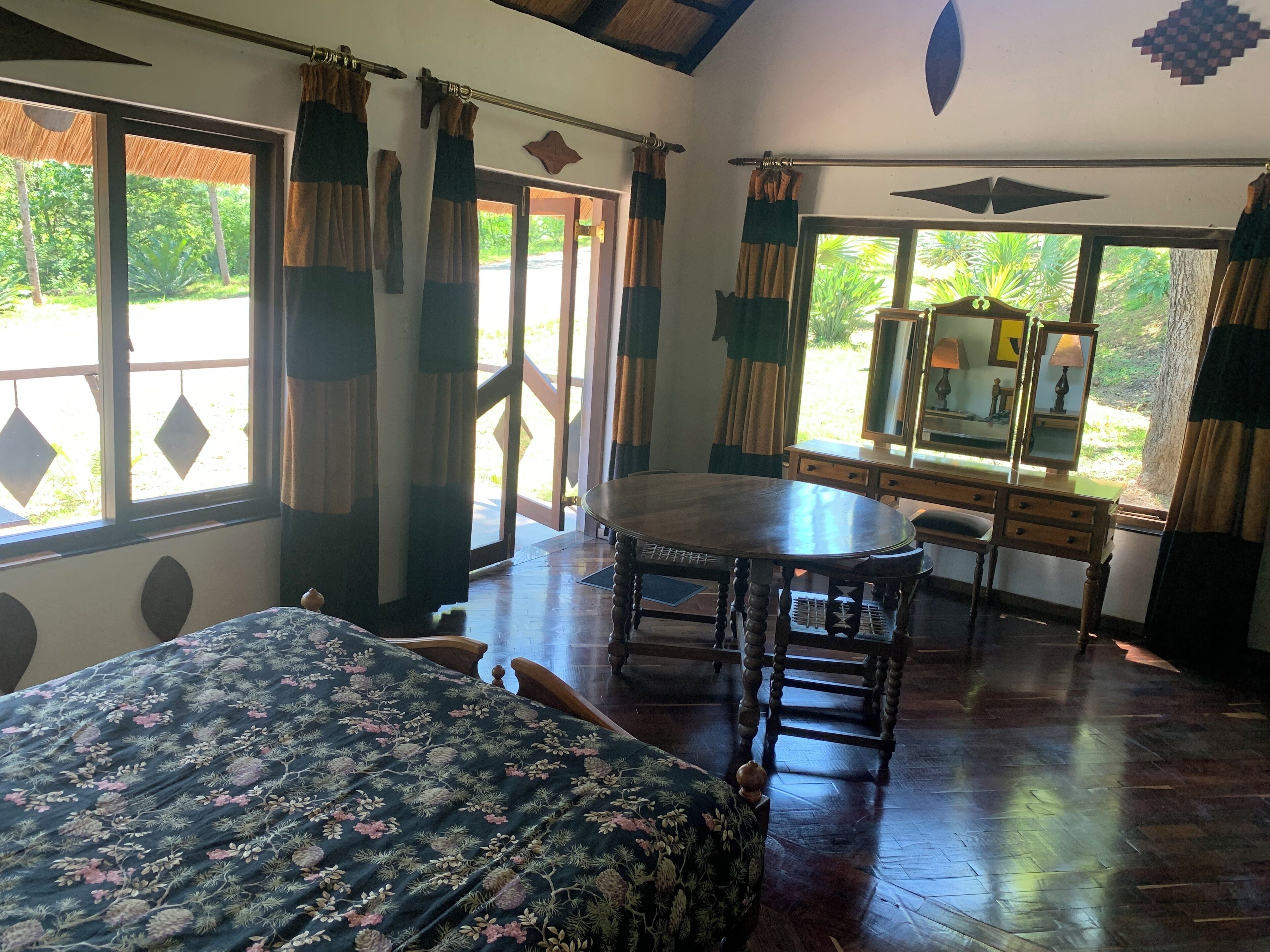 A room with a bed, a round wooden table, a desk with a mirror, and large windows with curtains.