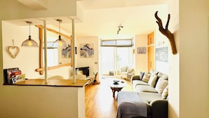A spacious living area with a wooden floor, a sofa, a coffee table, and a dining area with a table and chairs. There is a large window providing natural light and a view of the outdoors. - Luxury Seacliff Apartment overlooking Ventnor Beach, Sleeps 4 , Ferry Discounts (Ventnor)