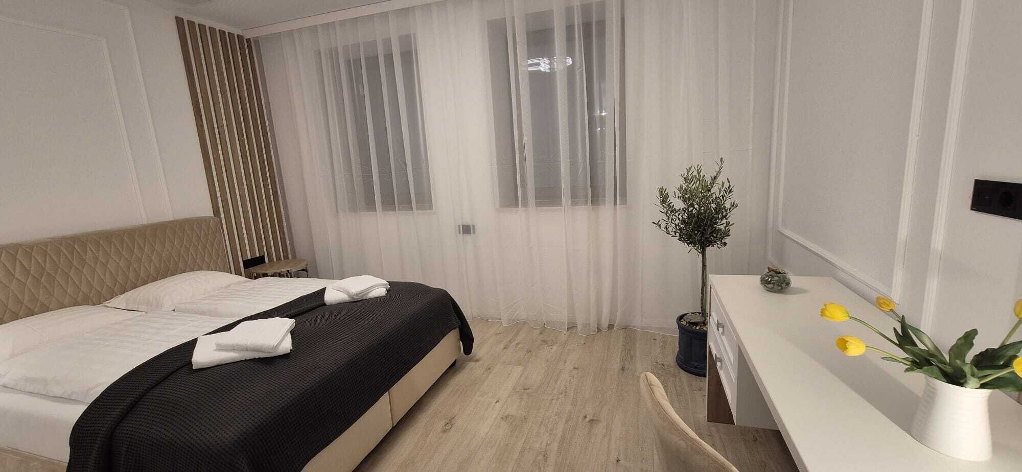 Ein modernes Hotelzimmer mit Bett, Nachttisch, Zimmerpflanze und einem Fenster mit durchsichtigen Vorhängen.