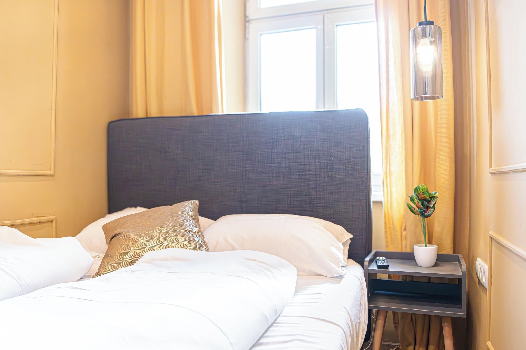 Een net opgemaakt bed met witte lakens en een grijs hoofdbord, een nachtkastje met een potplant en een hanglamp.