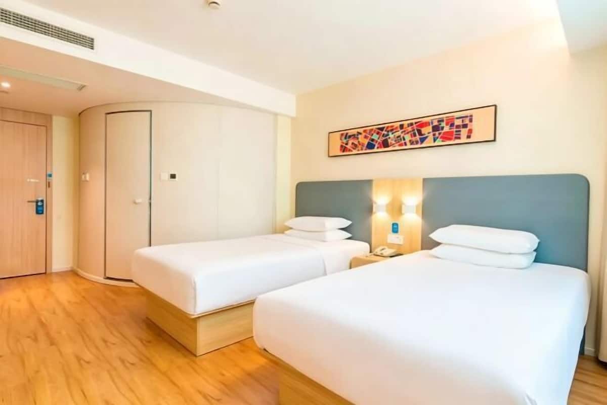 A hotel room with two beds, a wardrobe, a painting on the wall, and a nightstand with a lamp.
