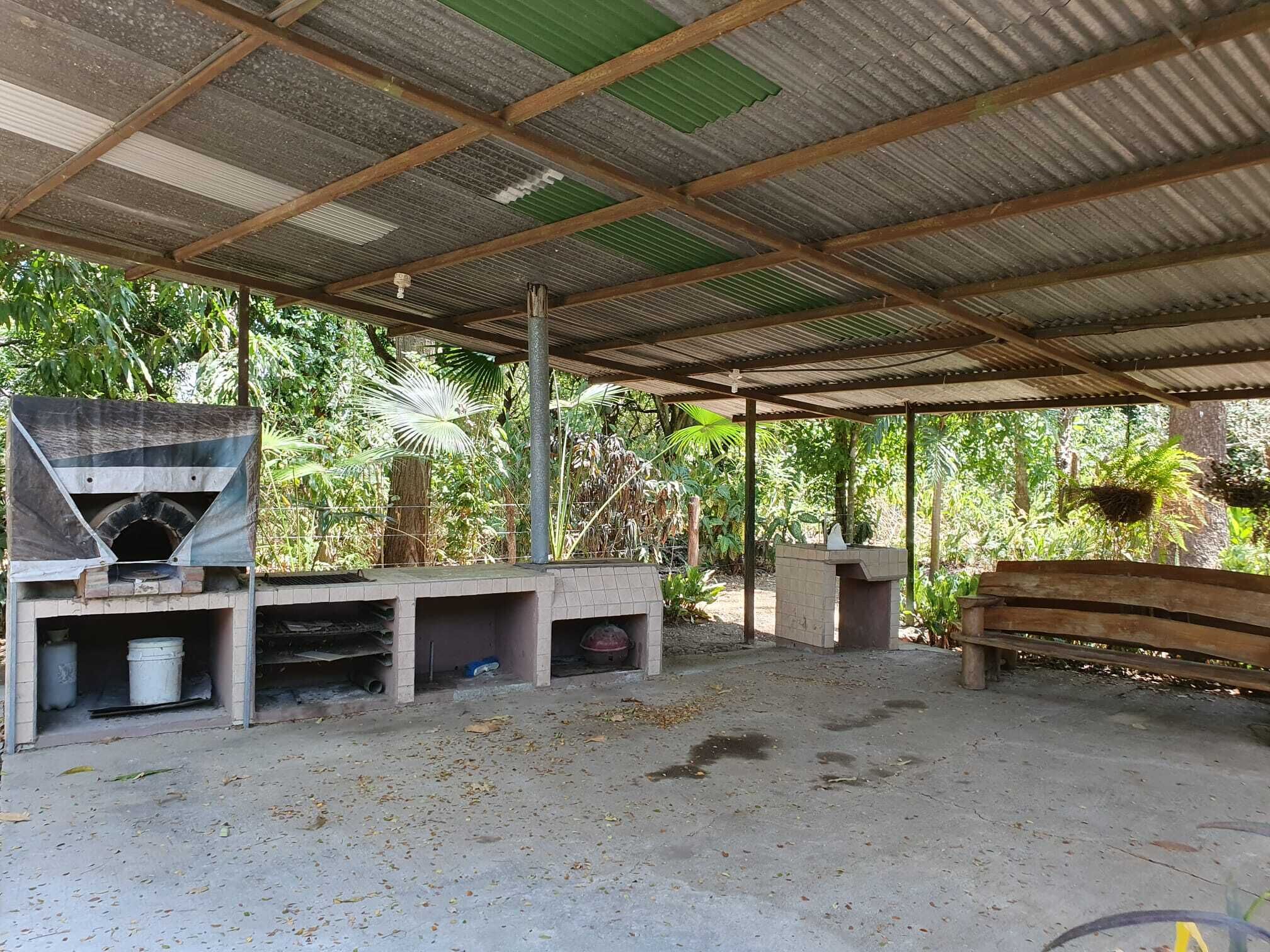 A rustic outdoor area with a brick oven, a wooden bench, and a covered shelter with a corrugated metal roof.