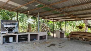 A rustic outdoor area with a brick oven, a wooden bench, and a covered shelter with a corrugated metal roof.