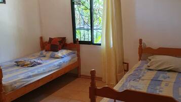 A room with two wooden beds, a window with a view of greenery, and a ceiling fan.