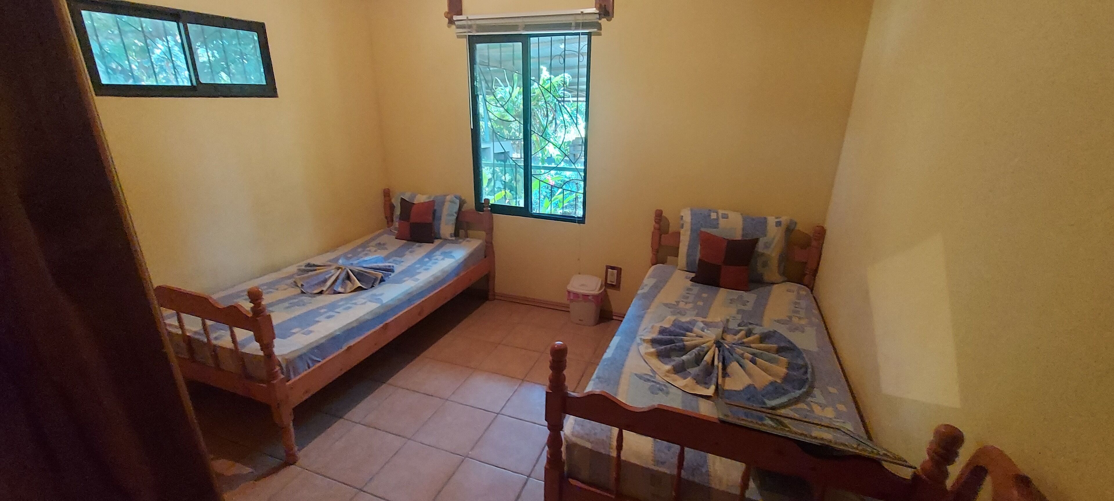 A small, compact room with two wooden beds, a window with bars, and a view of greenery outside.