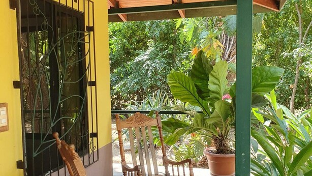 A covered patio with two wooden rocking chairs, a potted plant, and a window with metal bars.