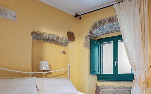 A bedroom with a bed, a lamp, a stone archway, a window with green shutters, and a curtain. - Sardinian Gallery Fonte (Bosa)