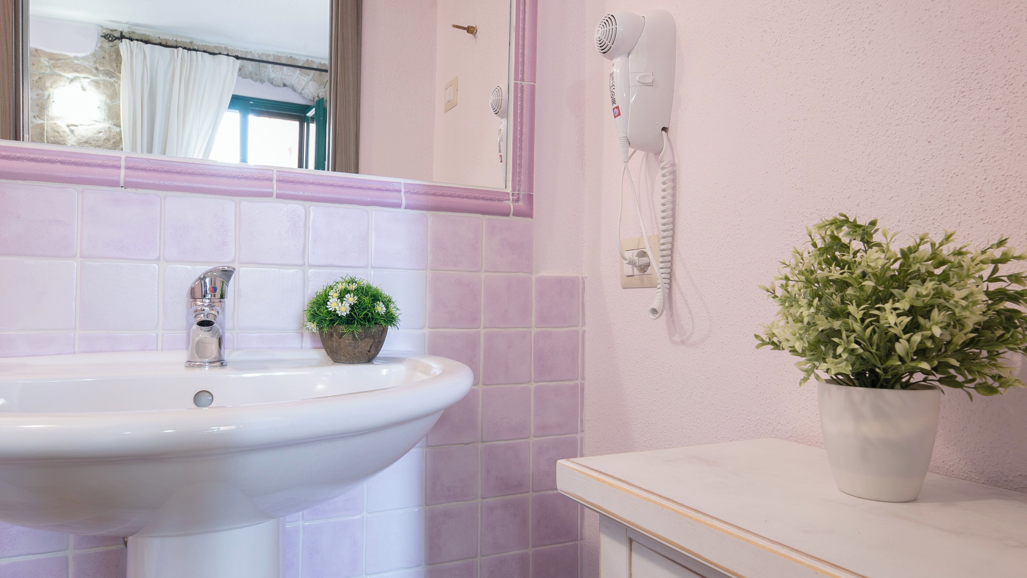 Un bagno con un lavandino bianco, una piccola pianta in vaso, un asciugacapelli e uno specchio.