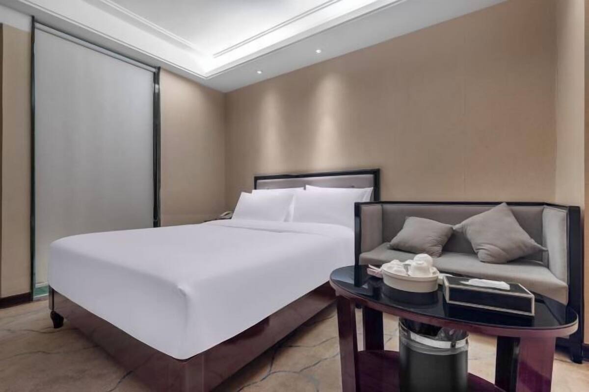 A hotel room with a bed, a sofa, a small table, and a trash bin.