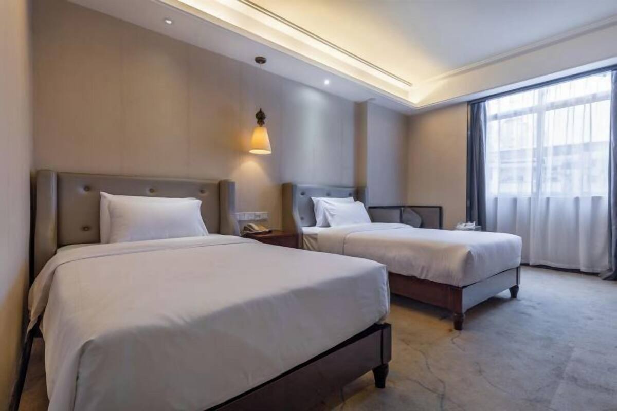 A hotel room with two beds, a nightstand, a lamp, and a large window with curtains.