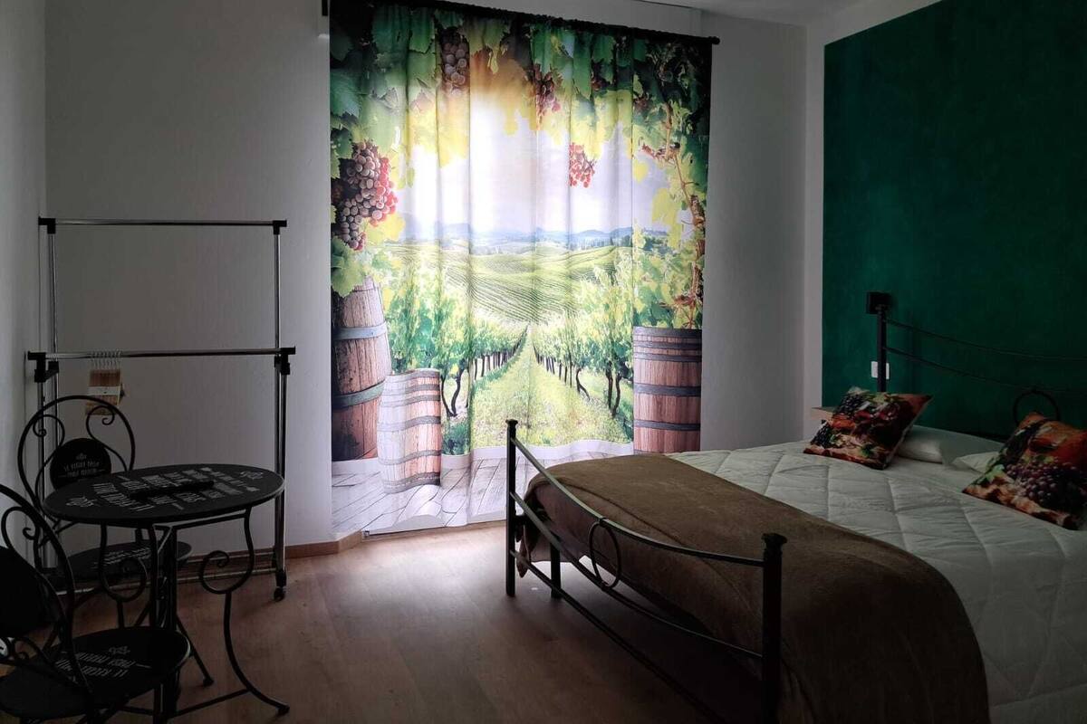 A bedroom with a bed, a green accent wall, a window with a vineyard scene curtain, a metal chair, and a small table.