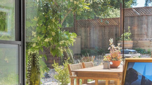 Een kamer met een groot raam dat uitkijkt op een tuin, een houten tafel met stoelen, een vaas en een lijst met een afbeelding.
