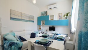 A modern living room with a white sofa, teal throw blanket, and a dining area with a white table set for four. - Galactic Sea: delightful cottage surrounded by greenery just minutes from the beach (Lido delle Nazioni)