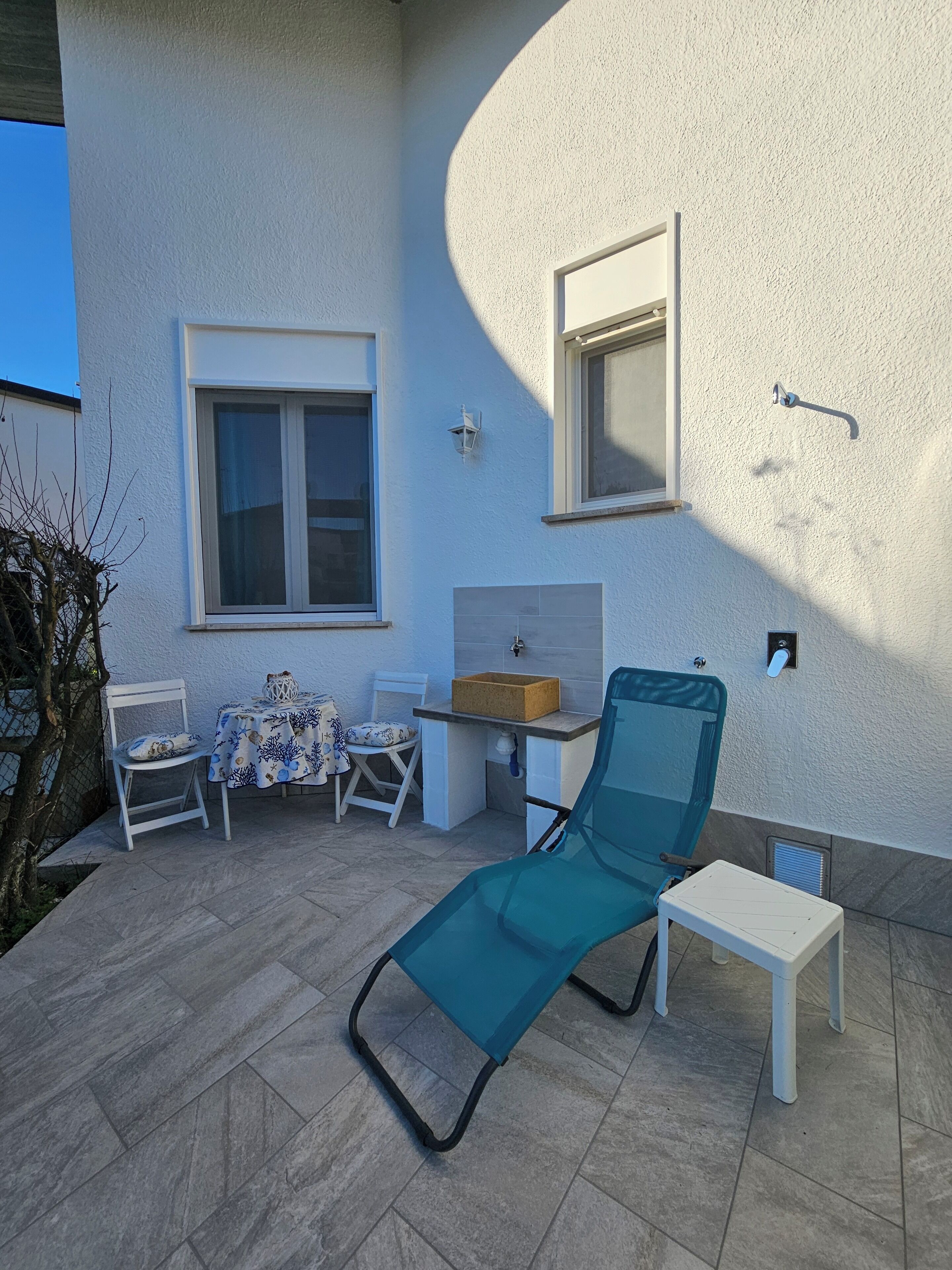 A patio area with a lounge chair, a small table, and a chair with a table. There is a window and a wall-mounted light fixture.