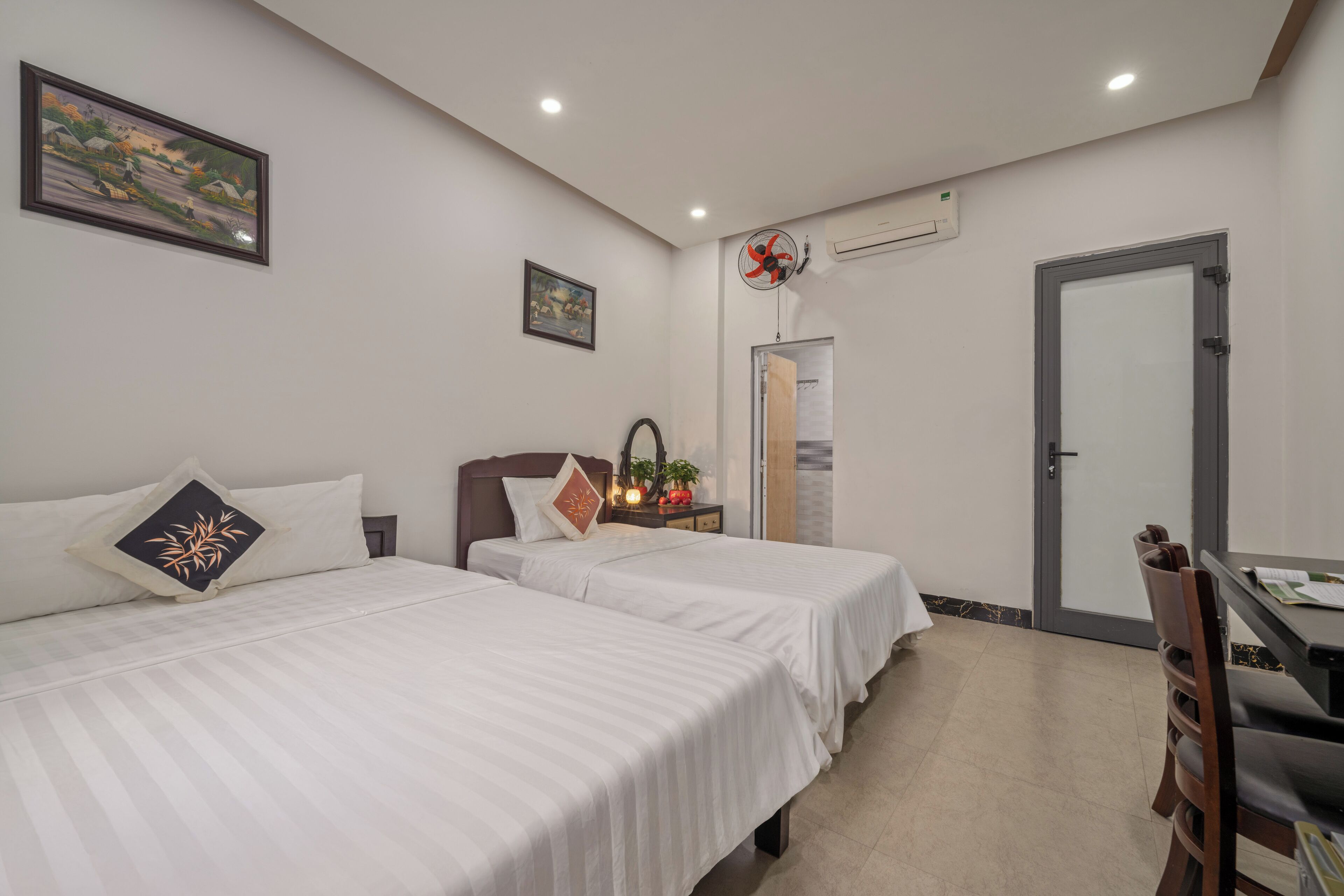 A hotel room with two beds, a dining table, and a chair. There is a door, a window, and a wall-mounted air conditioning unit.