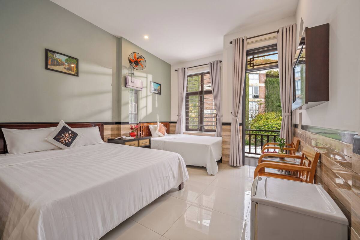 A hotel room with two beds, a balcony with wooden chairs, a TV, and a window with curtains.