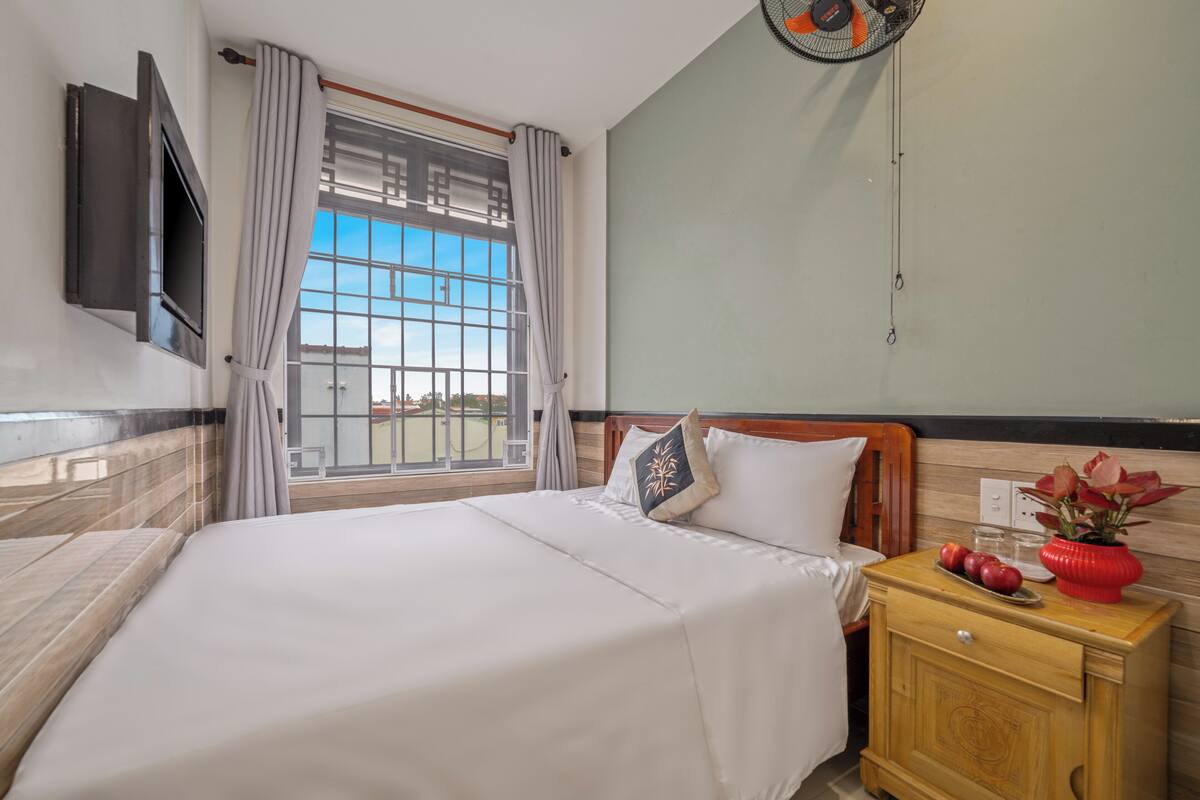 A hotel room with a bed, a bedside table with a vase and fruit, a TV mounted on the wall, and a window with curtains.