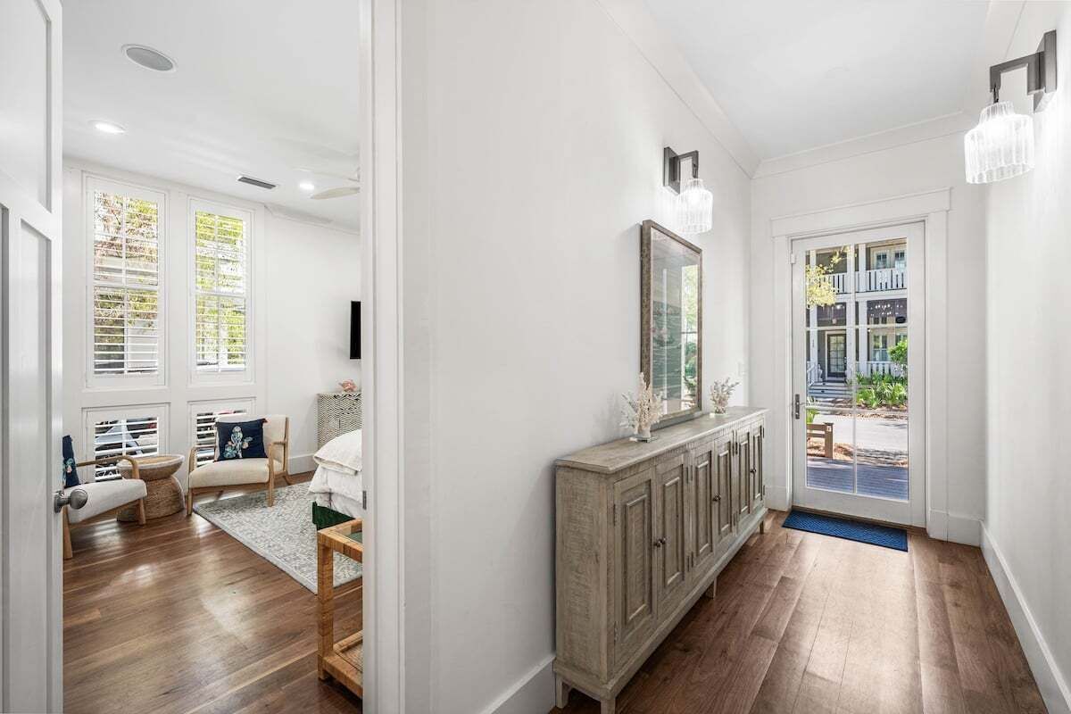 A bright hallway with wooden flooring, a white wall, a wooden console table with a mirror, and a door leading outside.