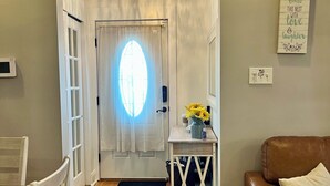 Ein Flur mit Holzboden, einer weißen Tür mit Fensterglas, einer Kandelaber, einem kleinen Tisch mit Blumenvase und einem braunen Ledersofa.