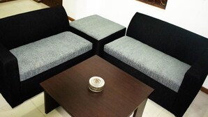 Une pièce comprenant deux canapés noirs avec des coussins gris, une petite table en bois ornée d’un objet décoratif et un sol carrelé blanc.